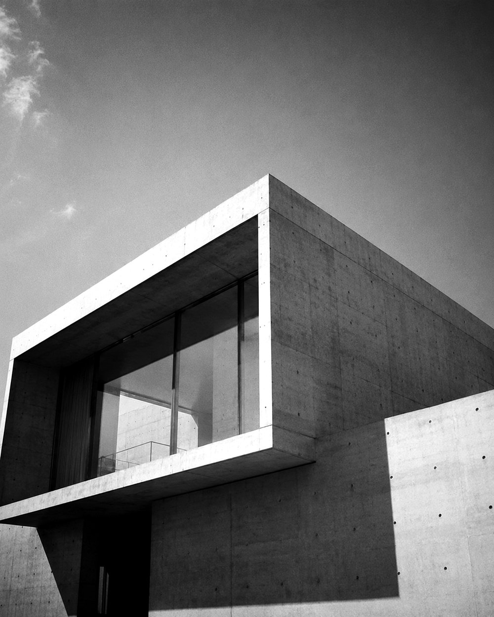 A modernist concrete house photographed from below in black and white — its cantilevered upper volume and glass corner window set against an open sky.