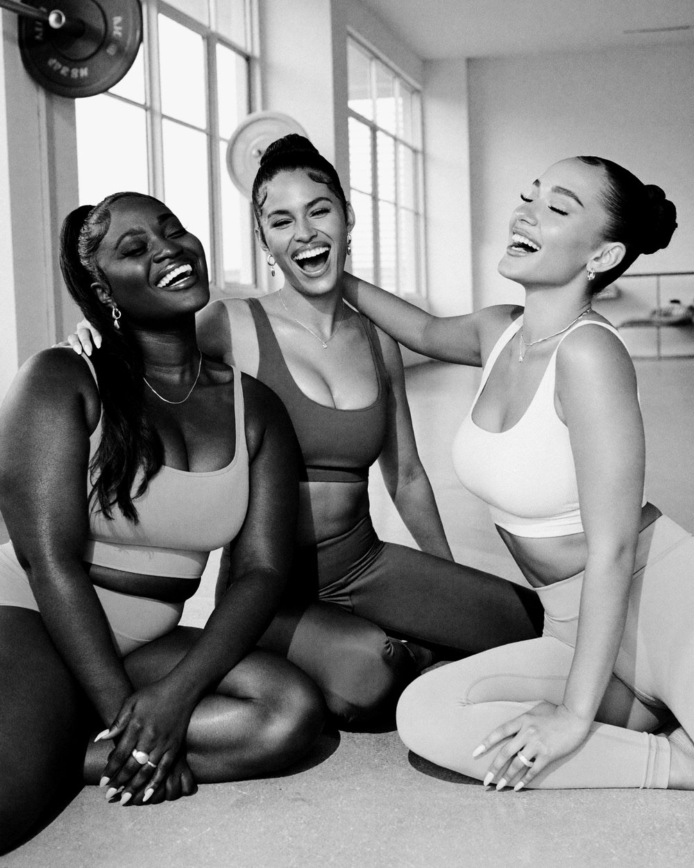 A bright black-and-white editorial photograph of three women laughing together in a sunlit gym, sitting close in matching athletic wear.