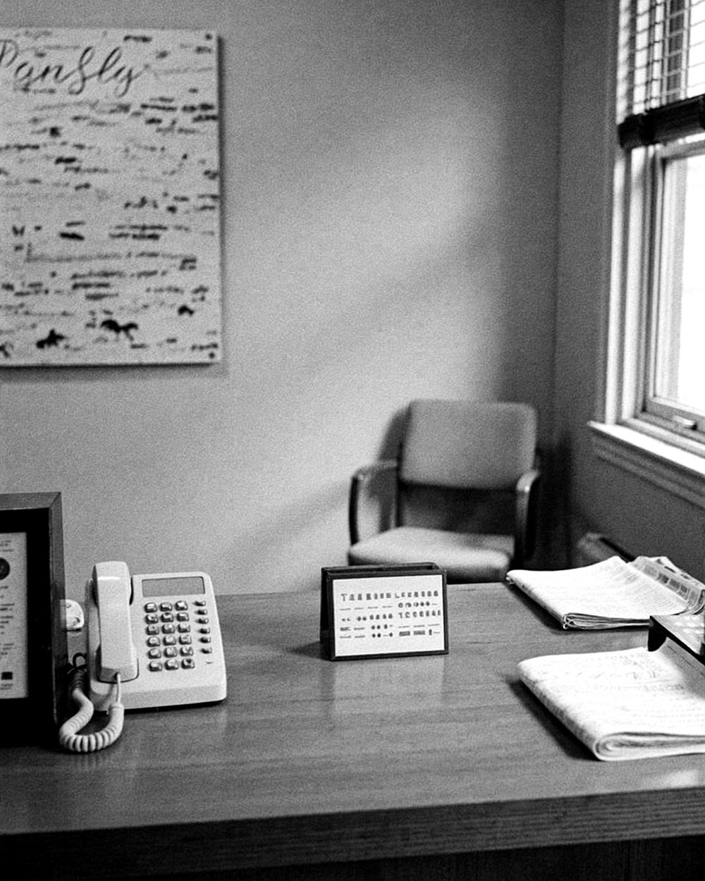 A quiet black-and-white photograph of a wood reception desk with a landline phone, a framed sign and a folded newspaper by a window.